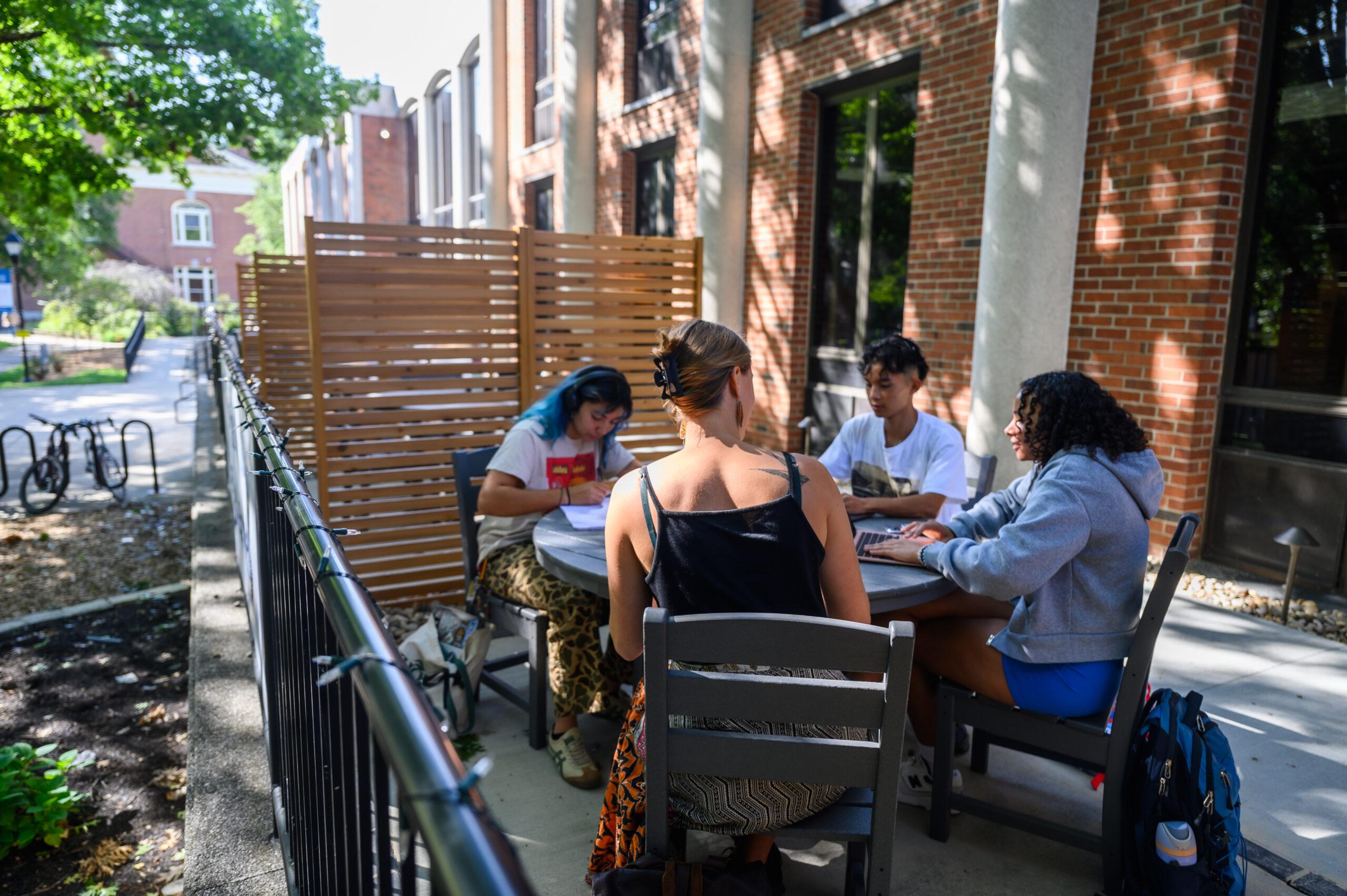 Diverse group of students seated at a table with a female instructor smiling as they lean between the students.