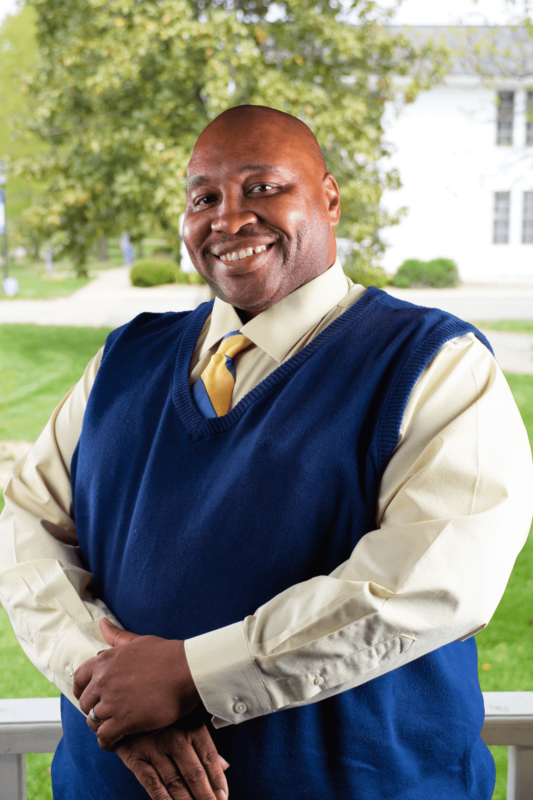 Portrait of Frank standing outdoors on campus, wearing a yellow button-down shirt, yellow tie, and navy sweater vest, with trees and a white building blurred in the background.