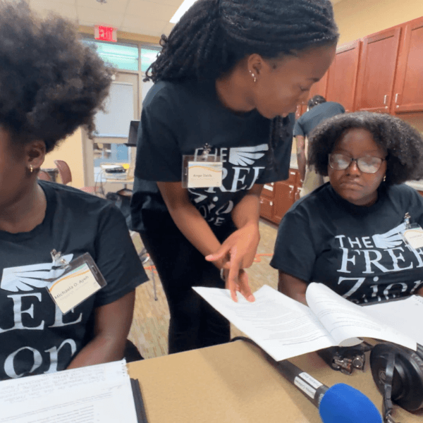 Three students sit behind a table and discuss while gesturing to paper documents.
