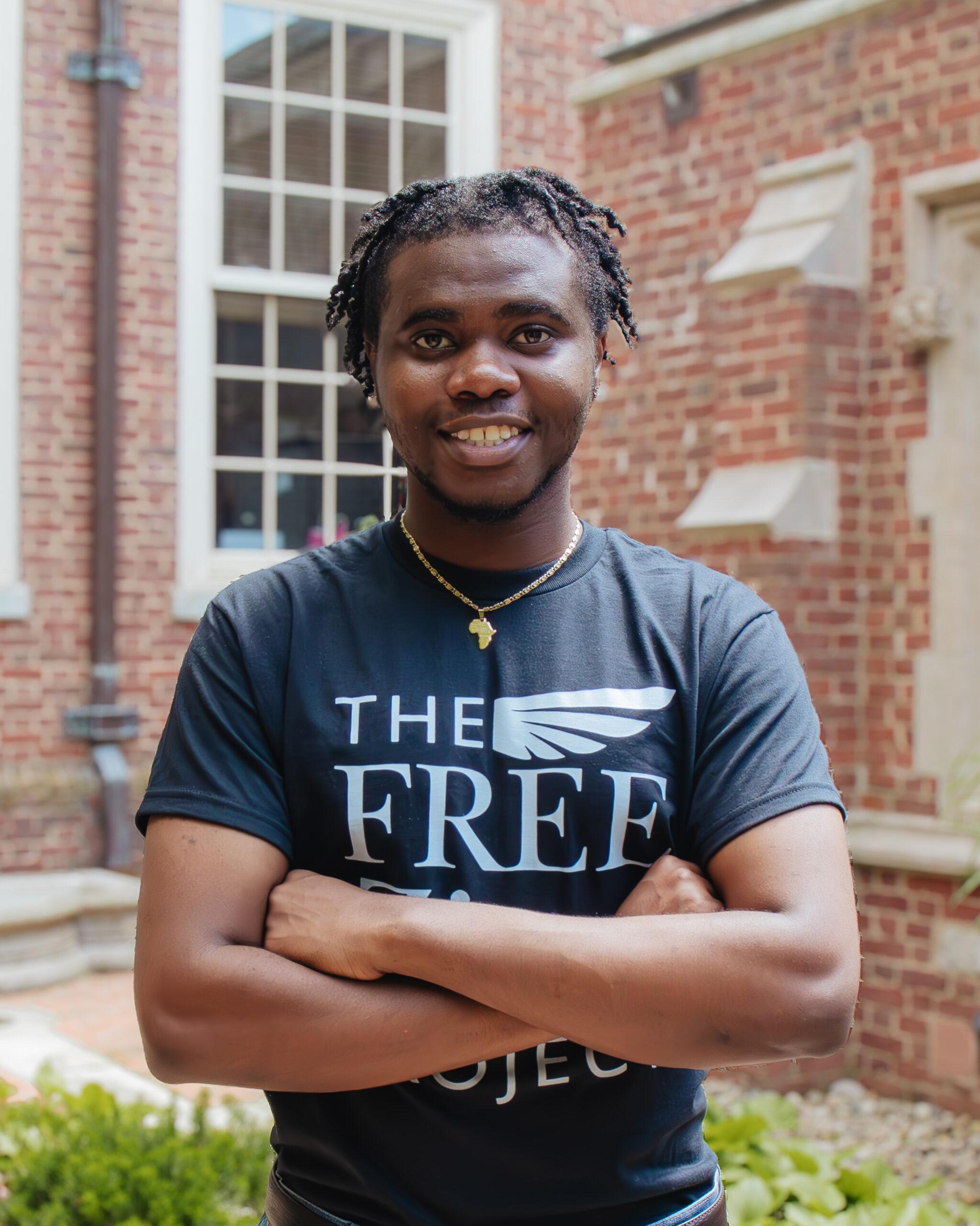 A male student wearing a black T-shirt smiles at the camera and crosses his arms in front of his chest.